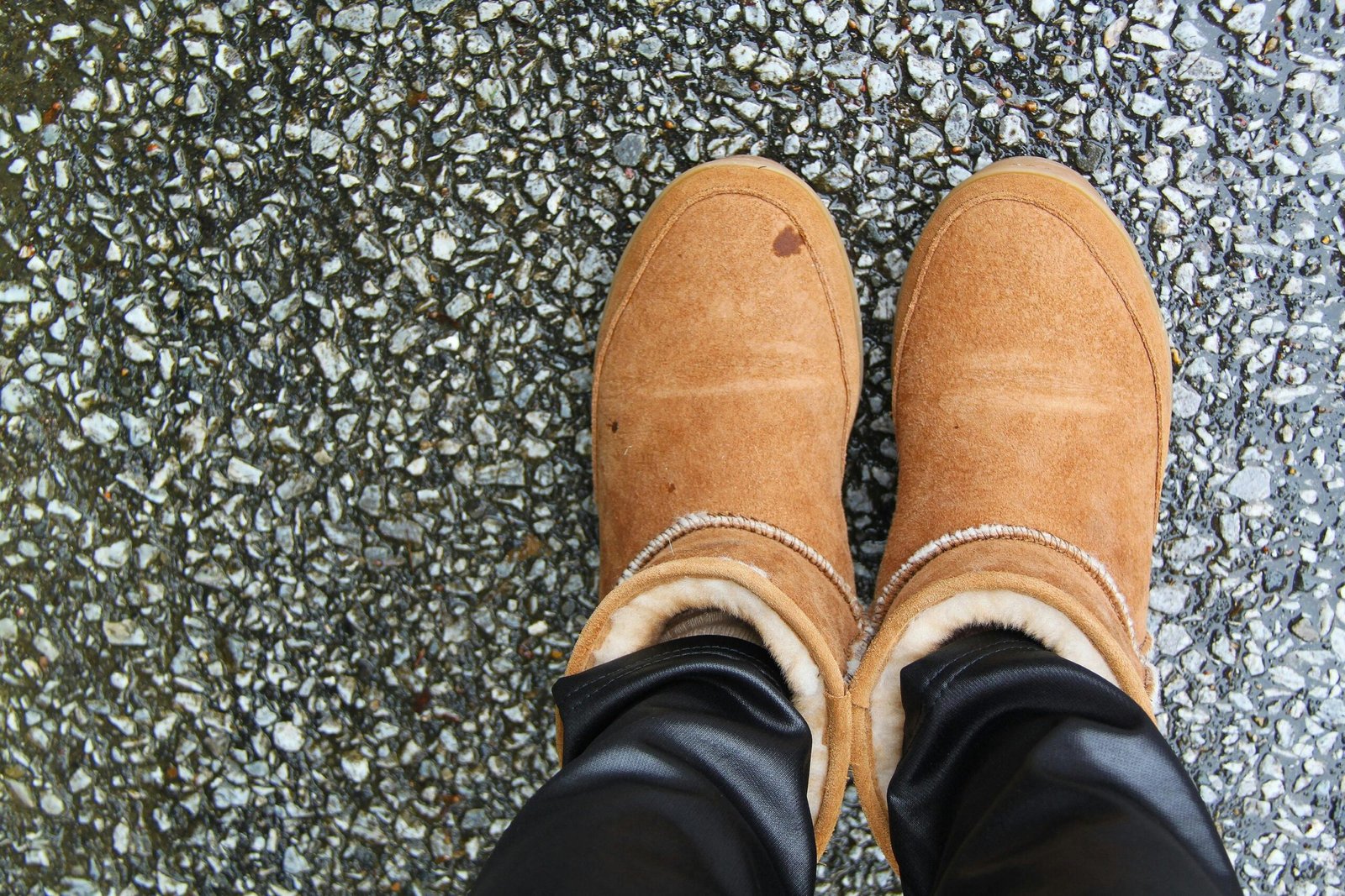 A pair of light brown suede winter boots with fur lining, worn by a person standing on wet asphalt.